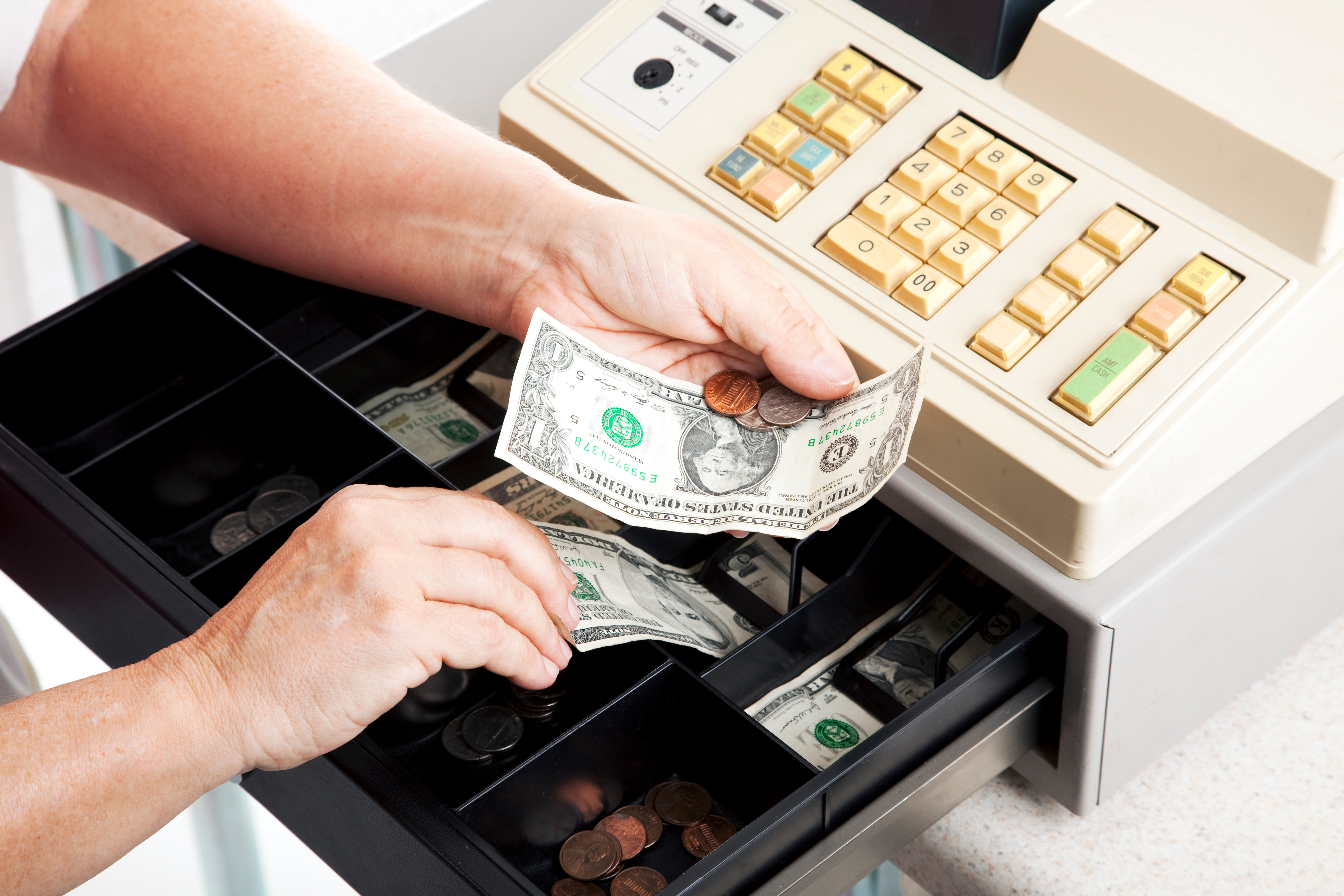A person taking bills (one dollar, five dollar, and a handful of coins) out of a cash register.
